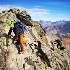 Wspinacze pokonują skalistą grań, a jeden z nich, w zielonym kasku, zmierza w stronę szczytu. Widok na Tatry zapiera dech w piersiach, a świnica przez zawrat wydaje się być na wyciągnięcie ręki.