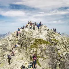 Tłum turystów na skalistym szczycie Rysów, z widokiem na Tatry. W dole majaczy Morskie Oko.
