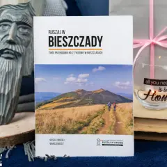Przewodnik "Ruszaj w Bieszczady" z informacjami o Bieszczadzkim Parku Narodowym. Na zdjęciu także rzeźba, szklanka z napisem i książki.