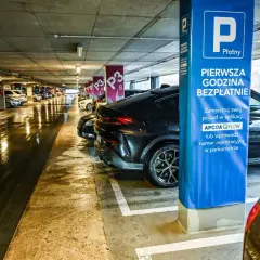 Mokry parking nad zaporą, rząd samochodów pod betonowym stropem. Niebieska tablica informuje o pierwszej darmowej godzinie.