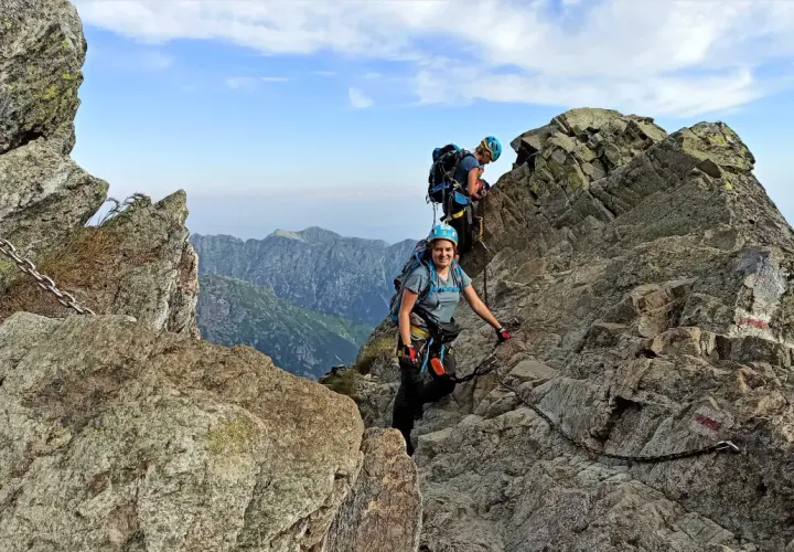 Dwoje turystów wspina się po skalistych tatry wysokie szlaki, korzystając z łańcuchów. Widok na góry i błękitne niebo.
