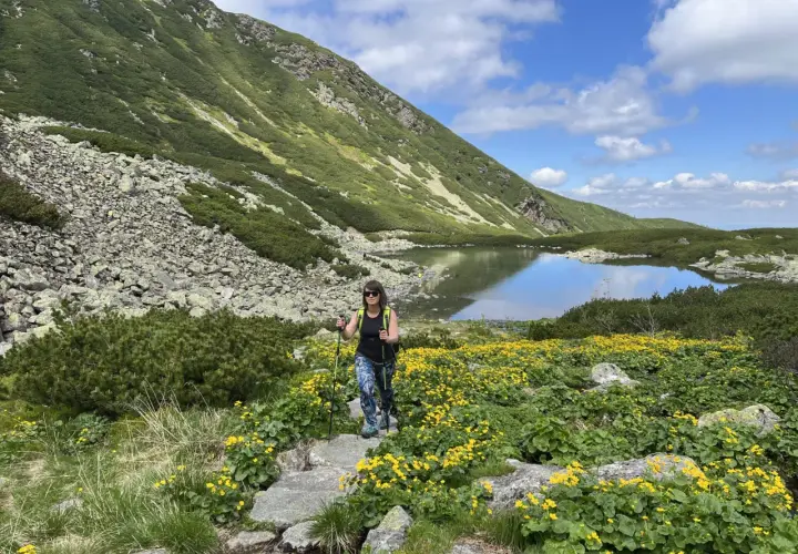 Kobieta z kijkami trekkingowymi idzie przez kwitnącą łąkę w górach. To najłatwiejszy szlak na Krzyżne, z jeziorem w tle.