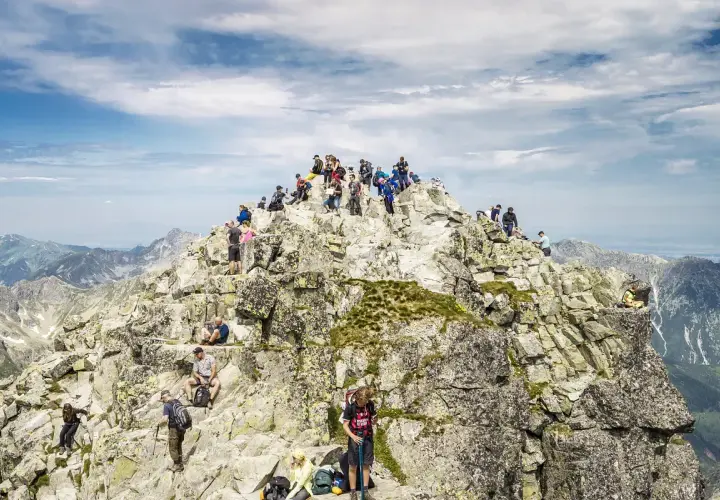 Tłum turystów na skalistym szczycie Rysów, z widokiem na Tatry. W dole majaczy Morskie Oko.
