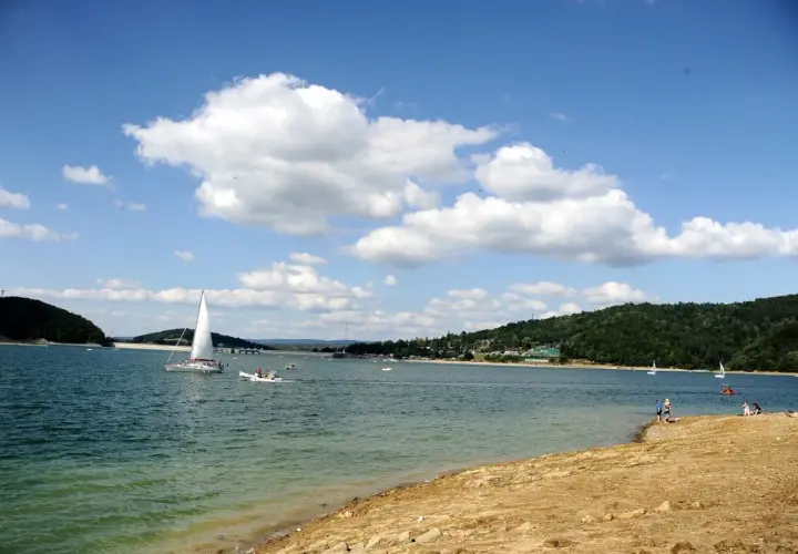 Słoneczna plaża nad Soliną, z żaglówkami na turkusowej wodzie i zielonymi wzgórzami w tle.
