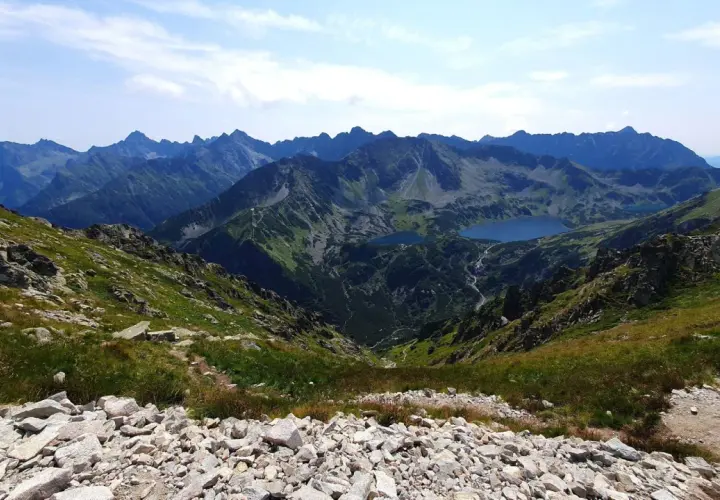 Widok na Tatry z kamienistego zbocza. W dole jeziora i kręte szlaki na krzyżne, otoczone zielenią i skalistymi szczytami.