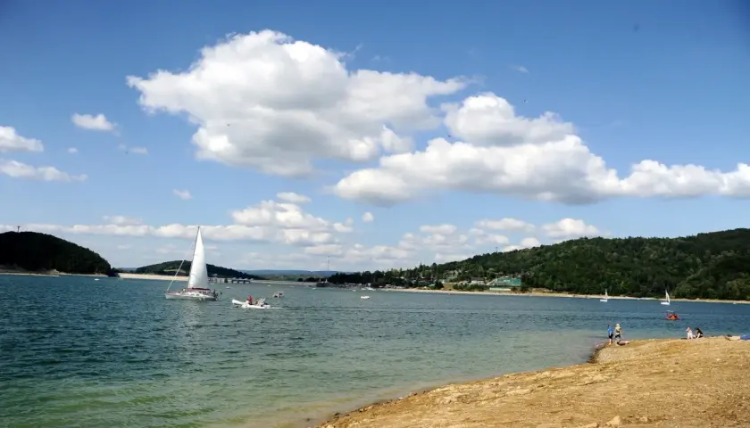 Słoneczna plaża nad Soliną, z żaglówkami na turkusowej wodzie i zielonymi wzgórzami w tle.