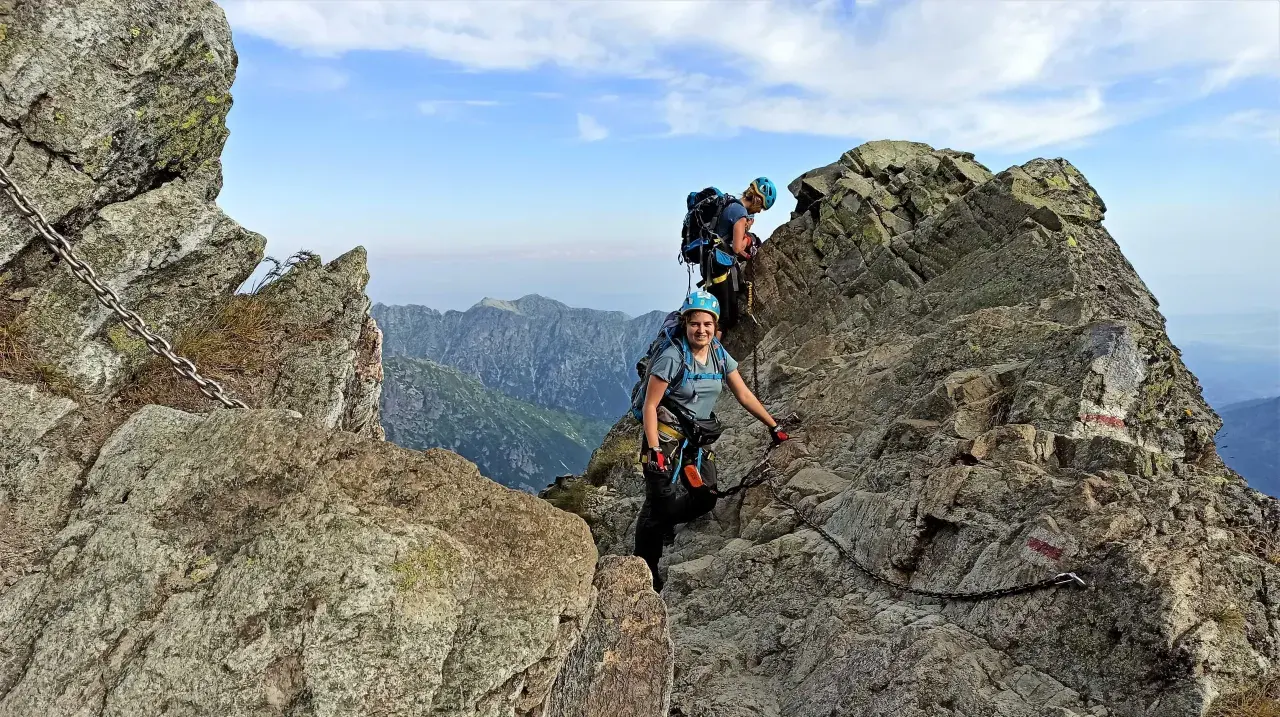 Dwoje turystów wspina się po skalistych tatry wysokie szlaki, korzystając z łańcuchów. Widok na góry i błękitne niebo.