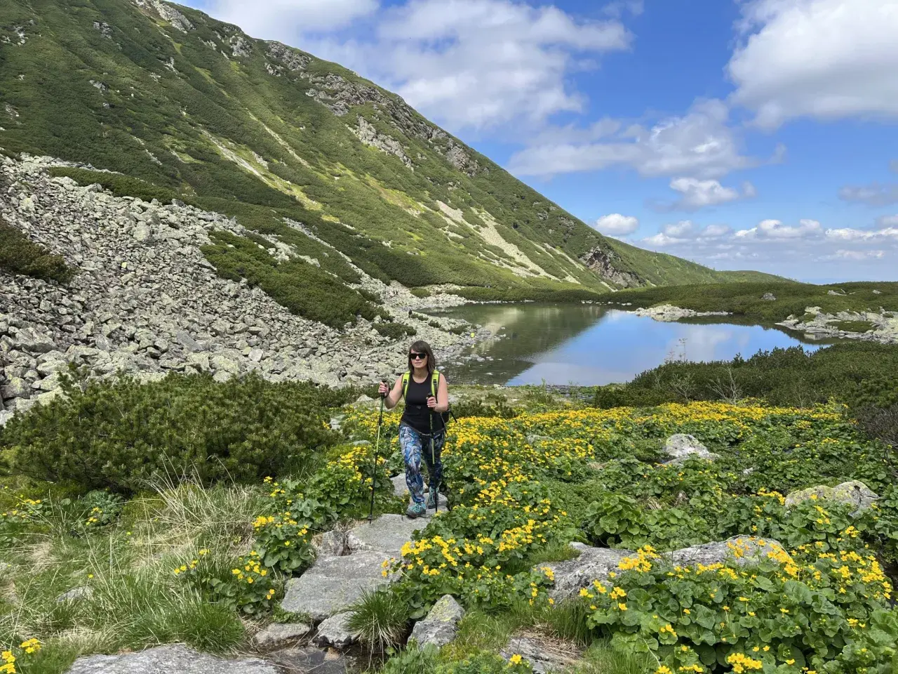 Kobieta z kijkami trekkingowymi idzie przez kwitnącą łąkę w górach. To najłatwiejszy szlak na Krzyżne, z jeziorem w tle.