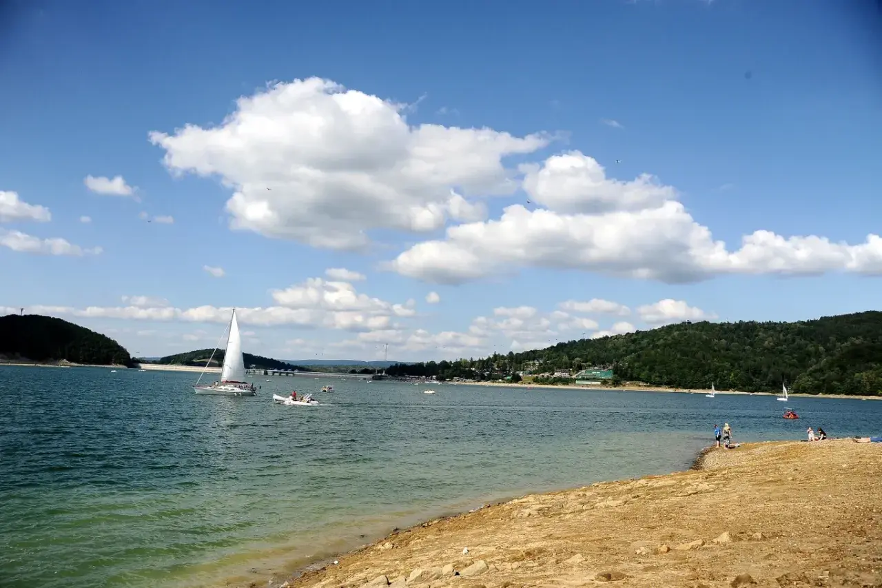 Słoneczna plaża nad Soliną, z żaglówkami na turkusowej wodzie i zielonymi wzgórzami w tle.