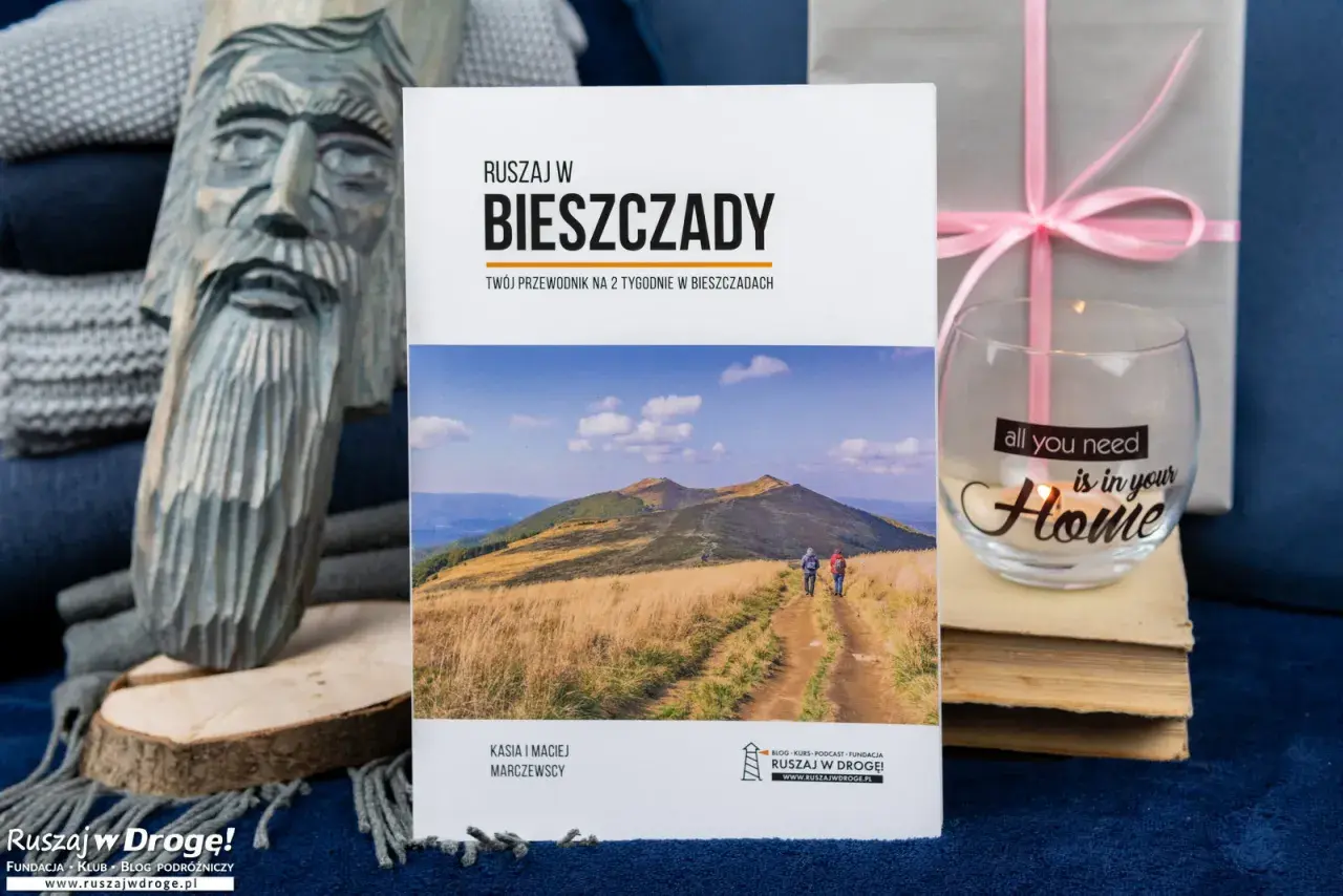 Przewodnik "Ruszaj w Bieszczady" z informacjami o Bieszczadzkim Parku Narodowym. Na zdjęciu także rzeźba, szklanka z napisem i książki.