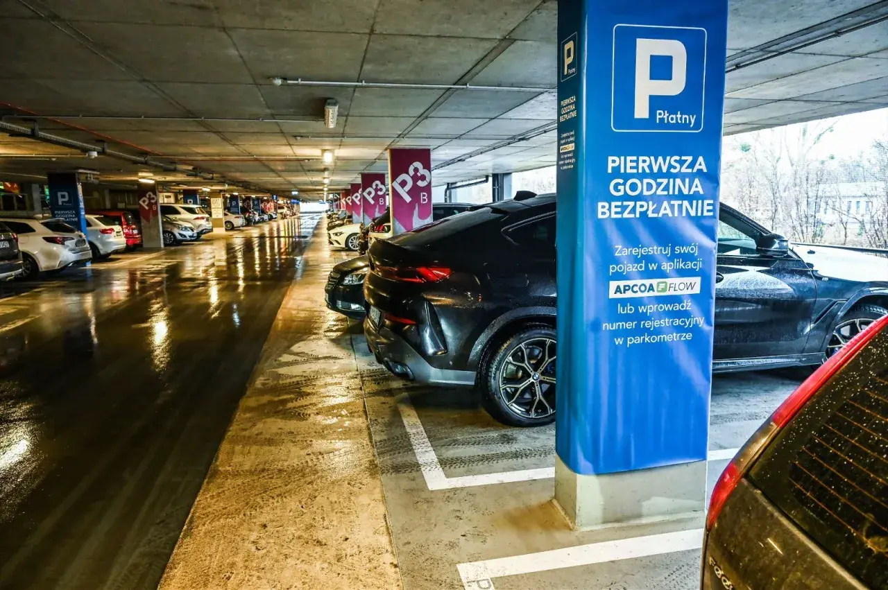 Mokry parking nad zaporą, rząd samochodów pod betonowym stropem. Niebieska tablica informuje o pierwszej darmowej godzinie.