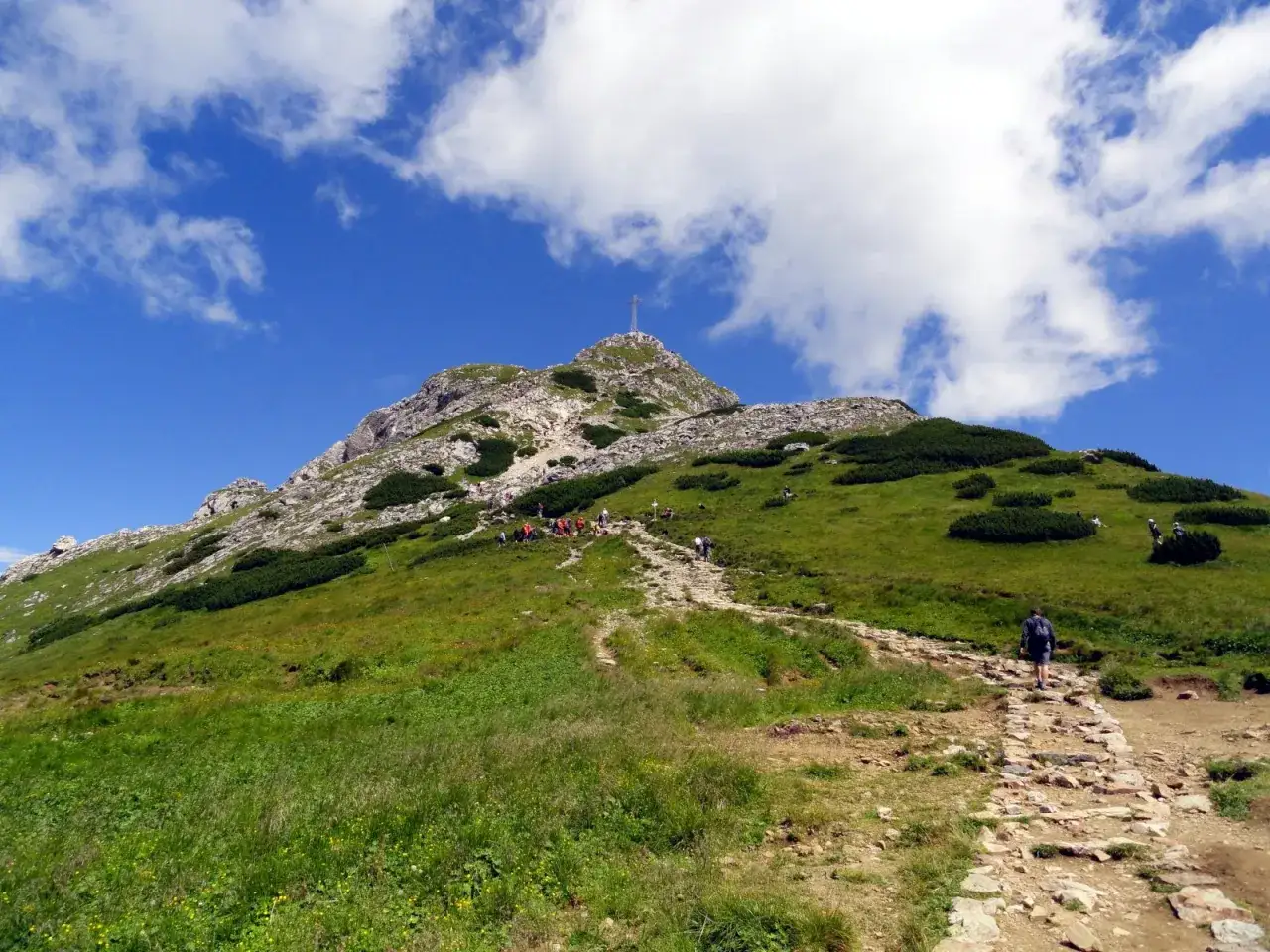 Giewont z Doliny Strążyskiej. Kamienista ścieżka wije się po zielonym zboczu, po którym wędrują turyści. Na szczycie widoczny krzyż.
