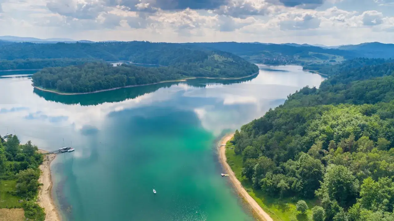 Malownicze jezioro w Bieszczadach otoczone zielonymi lasami, z kilkoma żaglówkami przy brzegu.