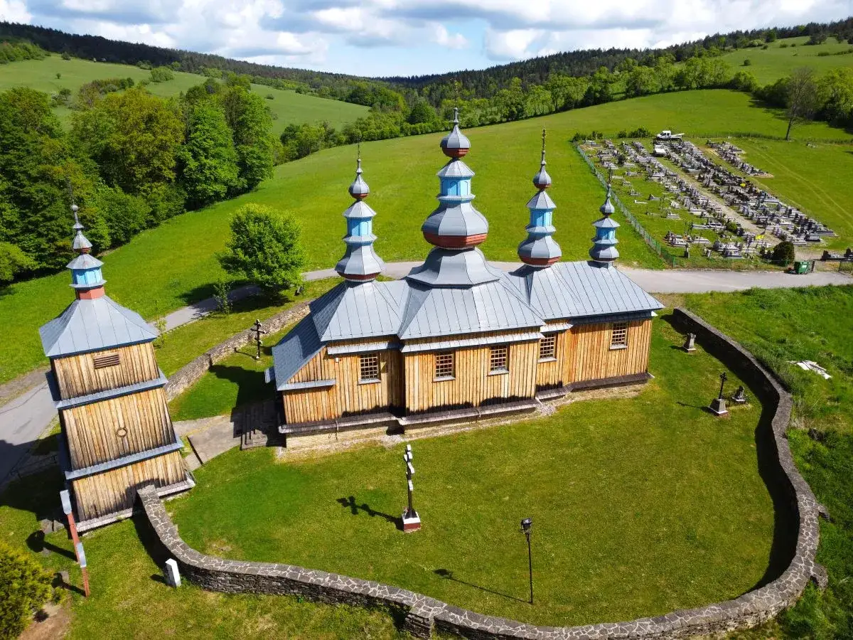 Drewniana cerkiew w Komańczy, z charakterystycznymi kopułami, otoczona zielonymi wzgórzami i cmentarzem.