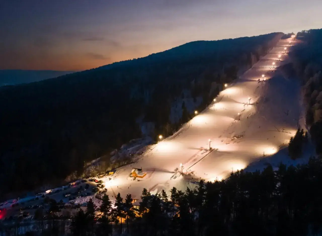 Oświetlony stok narciarski w Solinie nocą. Widoczne lampy, kolejka, budynki i zaparkowane samochody. Solina zimą atrakcje dla narciarzy.