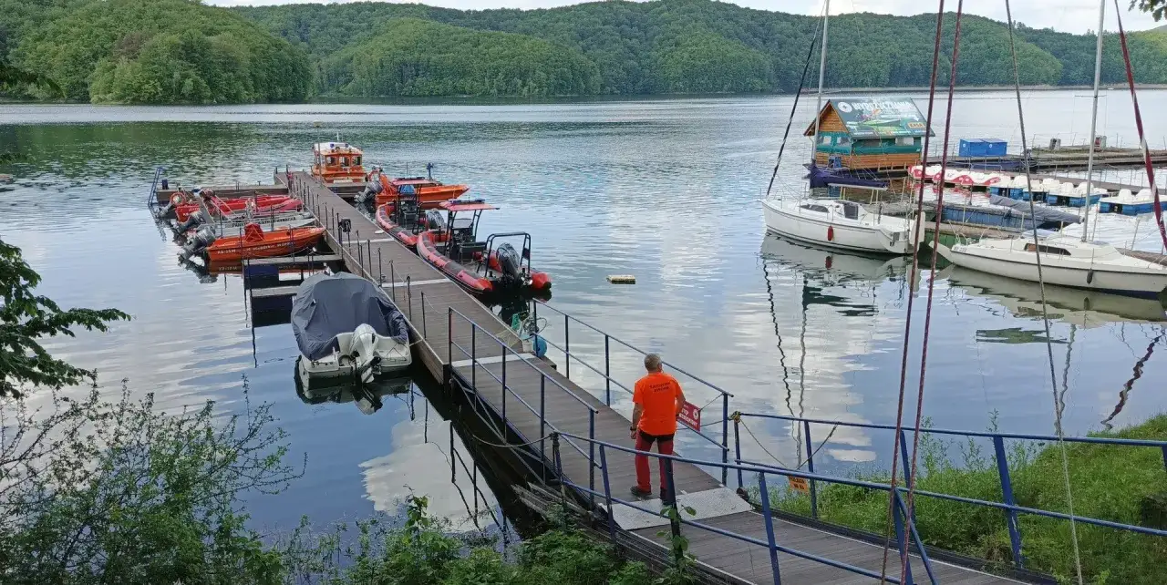 Przystań na plaży nad Soliną, z łodziami ratowniczymi i żaglówkami. Mężczyzna w pomarańczowej koszulce idzie po pomoście.